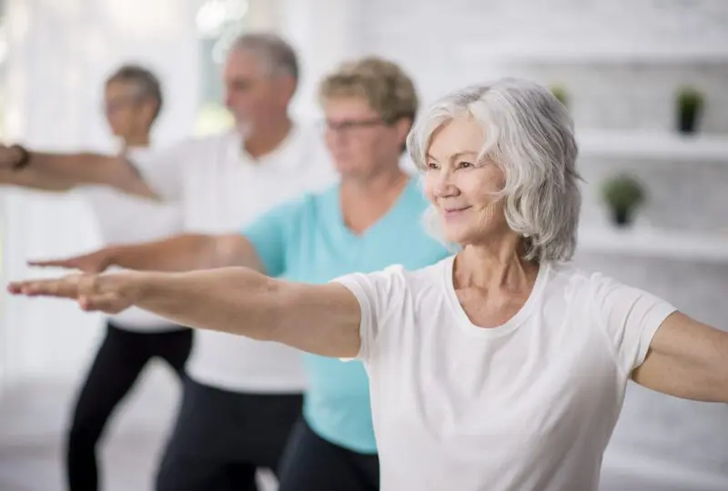 A multi-ethnic group of adult men and women are indoors in a fitness studio. They are wearing casual clothing while at a yoga class. A senior Caucasian woman is smiling while stretching out her arms.