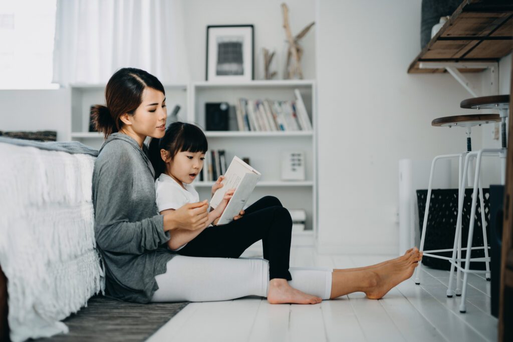 oung Asian mother sitting on the floor in the bedroom reading book to little daughter, enjoying family bonding time together at home