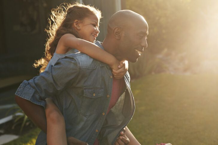a father smiling and running with his young daughter on his back.