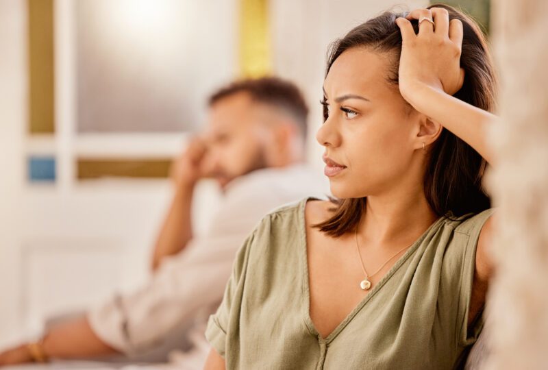 husband and wife seated away from eachother on couch, each looking concerned